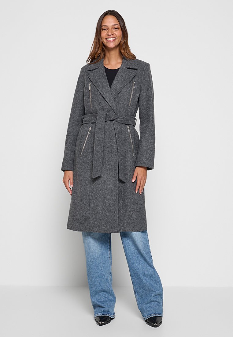 Femme souriante portant un manteau gris ceinturé avec des poches zippées, un haut noir, un jean large bleu et des chaussures noires à bout pointu, se tenant devant un fond blanc.