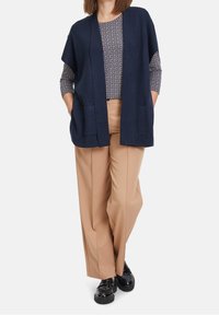 Marinblå cardigan med öppen front och sidfickor, lager-på-lager över en mönstrad topp; ihop med beiga vida byxor och glänsande svarta skor.