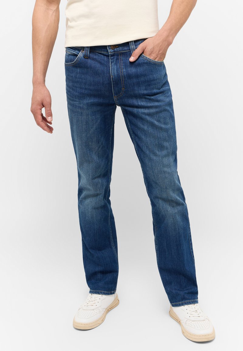 Herren Jeans aus blauen Denim mit geradem Schnitt, fünf Taschen, Kontrastnähten und einer natürlichen Ausbleichung. Getragen mit weißen Sneakers.