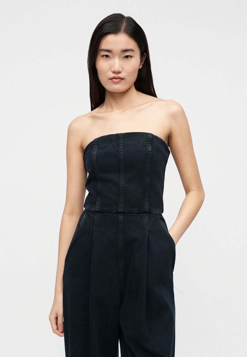 Combinaison noire en denim sans bretelles avec des détails de couture verticale, un design à jambes larges et des poches latérales. Texture mate avec un léger éclat.