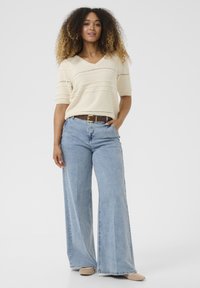 Maglione beige in maglia con scollo a V e maniche corte, abbinato a jeans in denim azzurro chiaro a gamba larga. Una cintura marrone mette in risalto il punto vita.