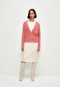 Cardigan rosa morbido con chiusura a doppio bottone e due tasche sul petto, abbinato a una gonna a tubino crema e stivali beige al ginocchio.