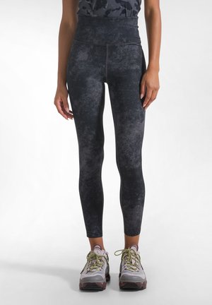 Kvinde iført sorte leggings med høj talje med et falmet mønster og lyse sportssko med grønne snørebånd, der står på en ensfarvet hvid baggrund.