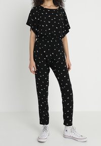 Svart jumpsuit med vitt fågelmönster, korta utblommade ärmar, elastisk midja och avsmalnande ben. Matchad med vita sneakers.