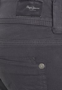 Jeans cinzentos escuros com detalhes cosidos, um pequeno bolso frontal e um botão de logotipo prateado na cintura. Etiqueta da marca visível na parte superior.