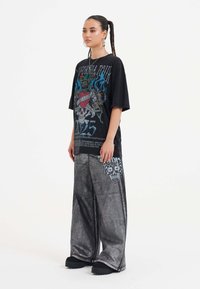 Černé oversized tričko s barevným grafickým designem; šedé, lesklé široké kalhoty se vzorem lebek; doplněné černými platformovými botami.