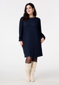 Marineblauwe gebreide jurk met lange mouwen, ontspannen pasvorm en zijsplitten, gecombineerd met crèmekleurige slouchy laarzen. Eenvoudig en veelzijdig ontwerp.