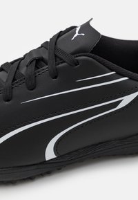 Sort sportsko med hvidt, buet stribedesign, sorte snørebånd og Puma-logo på tungen, vist i nærbillede på hvid baggrund.