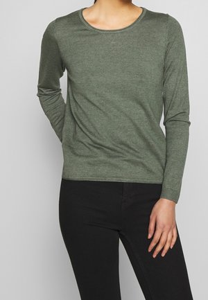 Person iført en tætsiddende olivengrøn langærmet skjorte og sorte bukser, stående mod en ensfarvet lysegrå baggrund.