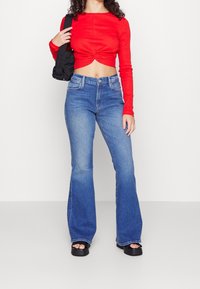 Top corto rojo acanalado con detalle de torsión, combinado con jeans acampanados de color azul claro y sandalias de plataforma negras. Los accesorios incluyen un bolso negro.