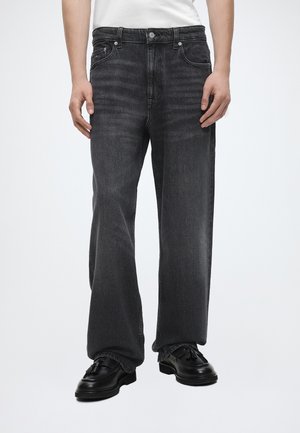 Person, der bærer falmede sorte jeans med lige ben og sorte loafers med kvaster, stående mod en ensfarvet lys baggrund.
