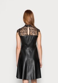 Schwarzes Lederkleid mit einem taillierten Oberteil und einem ausgestellten Rock. Spitze Details an den Schultern und am Rücken mit einem Schlüsselloch-Ausschnitt. Glatte Textur.