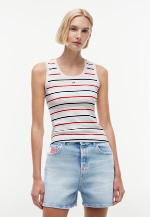 Femme aux cheveux blonds courts portant un haut blanc sans manches à rayures rouges et noires et un short en jean bleu clair.