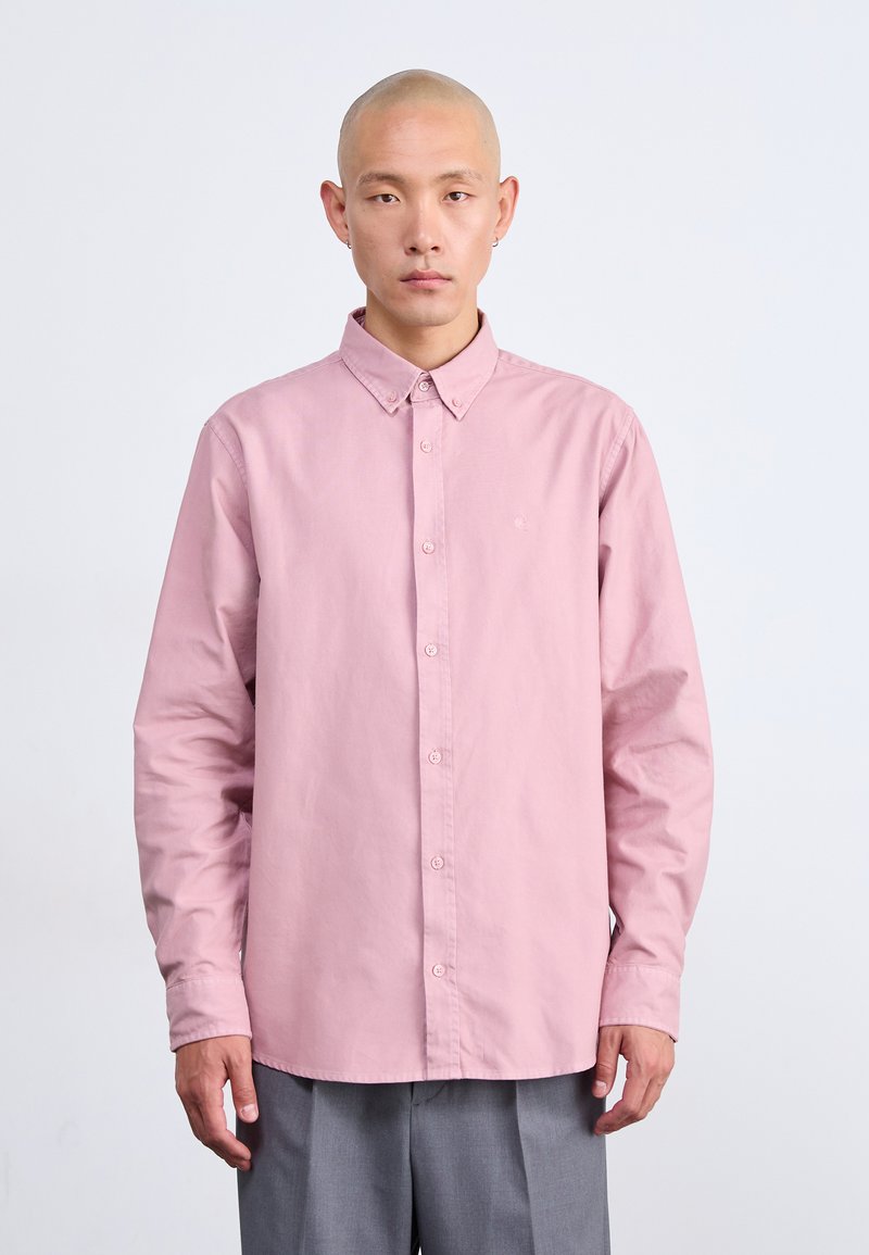 Camicia a maniche lunghe rosa chiaro con colletto, dotata di un logo discreto sul petto e vestibilità regolare, abbinata a pantaloni grigi.