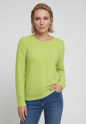 Frau mit kurzen blonden Haaren trägt einen limettengrünen strukturierten Langarmpullover und blaue Jeans, eine Hand in der Tasche.