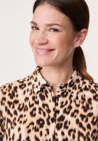 Tigreprint skjorte i tan og sort, med krave og knapper. Stoffet har en glat tekstur med en semi-gennemsigtig udseende.