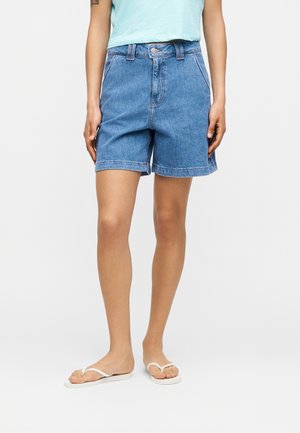 Personne portant un short en jean bleu mi-long, un haut sans manches bleu clair et des tongs blanches, debout contre un fond blanc uni.