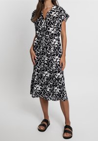 Schwarz-weißes, florales Midikleid mit kurzen Ärmeln, Knopfverschluss und Bindegürtel. Getragen mit schwarzen Hausschuhsandalen.