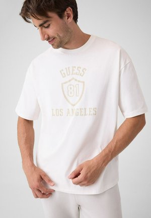 Mann trägt ein weißes Guess T-Shirt mit beigem Text und Schild-Logo mit der Aufschrift "81" und "Los Angeles,", schaut nach unten und lächelt leicht.