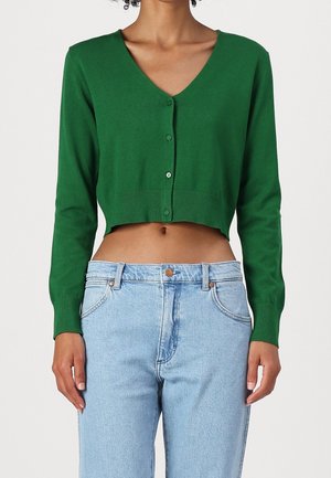 Femme portant un cardigan vert court boutonné à manches longues et un jean taille haute bleu clair, debout devant un fond blanc.
