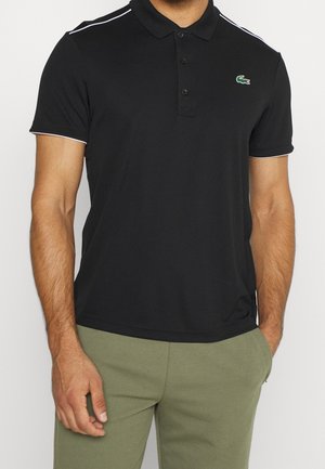 Man draagt een nauwsluitend zwart poloshirt met witte bies op de schouders en mouwen, gecombineerd met licht olijfgroene broek.