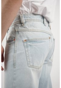Vaqueros de denim de color azul claro con cinco bolsillos, remaches de cobre y costuras en contraste. La tela tiene un aspecto ligeramente texturizado.