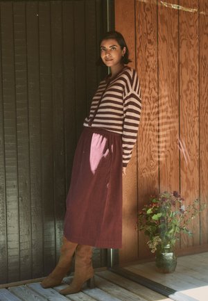 Femme portant un pull rayé, une longue jupe violette et des bottes marron, appuyée contre un mur en bois avec un vase en verre de fleurs à proximité.