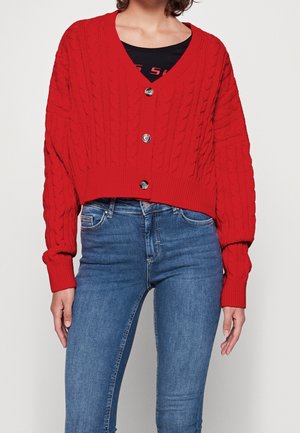 Rød kabelstrikket cardigan med knapper over sort top, kombineret med højtaljede blå skinny jeans på en stående person.