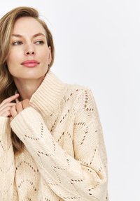 Beżowy sweter z dzianiny z prążkowanym golfem, z chevronowym wzorem ażurowym i szerokimi rękawami, prezentujący miękką, fakturowaną powierzchnię.