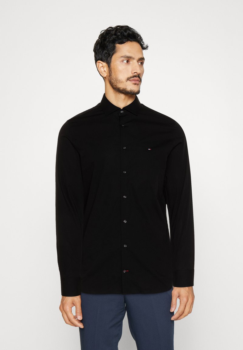 Schwarzes Langarmhemd mit Button-Down-Kragen, das ein tailliertes Design, eine glatte Textur und ein kleines, auf der Brust gesticktes Logo aufweist.