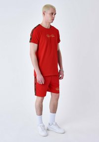 Conjunto de camiseta de algodón roja y pantalones cortos con logotipo en acentos negros y dorados en las mangas y los pantalones. El modelo lleva zapatillas y calcetines blancos.