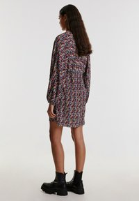 Blumenkleid mit langen Ärmeln und geraffter Textur, das ein lebhaftes Muster aus roten, blauen und gelben Blumen auf dunklem Hintergrund zeigt. Schwarze Knöchelstiefel.