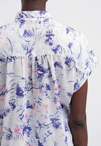 Personne portant une chemise blanche à manches courtes avec des motifs floraux bleus et roses, vue de dos sur un fond uni.