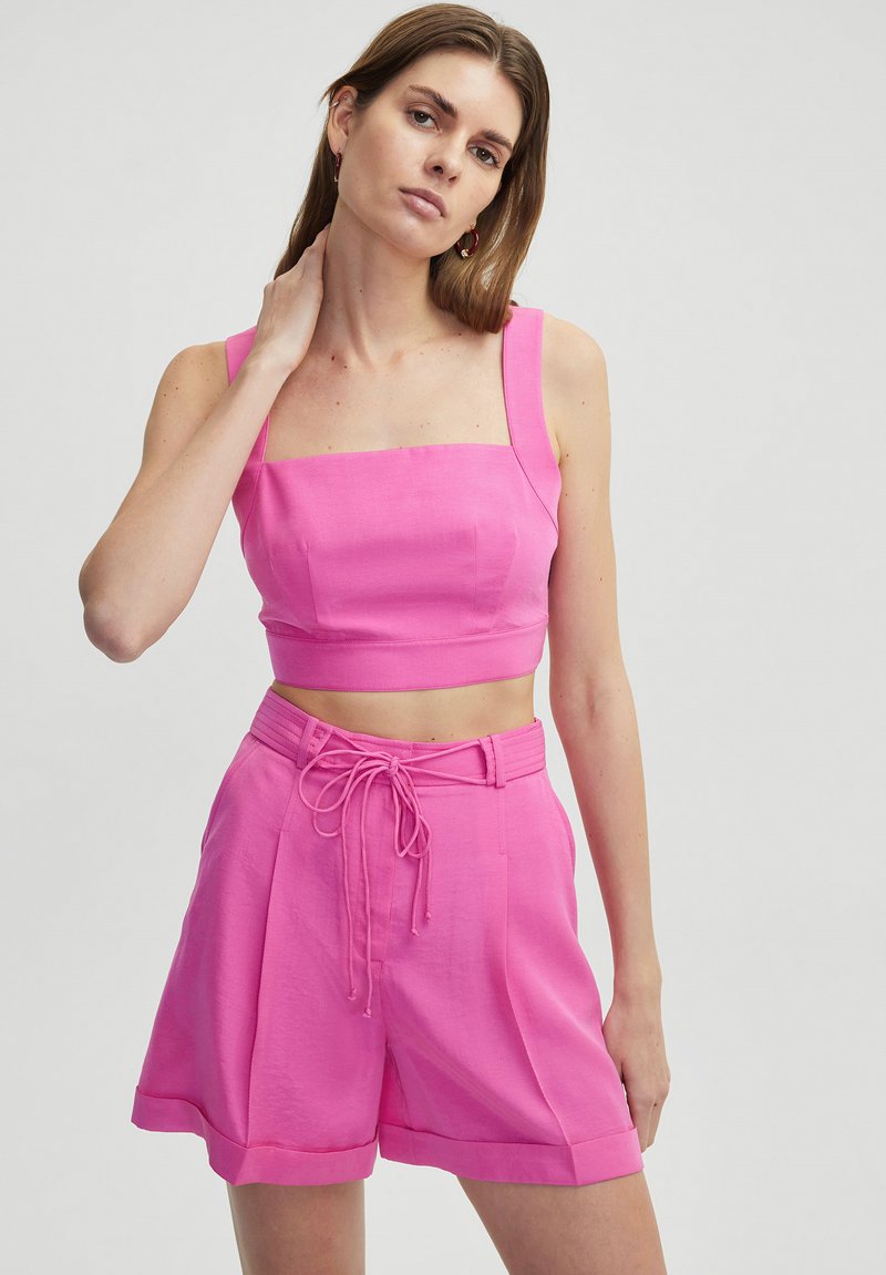Top corto rosa con escote cuadrado y pantalones cortos de cintura alta a juego. La tela tiene una textura suave, con un detalle de lazo en el frente y pliegues.