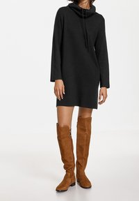 Robe en tricot noire avec encolure drapée et manches longues, accompagnée de bottes en daim marron jusqu'au genou avec zips sur les côtés.