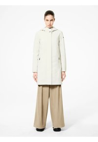 Cappotto impermeabile beige chiaro con chiusura a zip, maniche lunghe, cappuccio e tasche laterali, indossato su pantaloni beige larghi. Texture liscia e opaca.