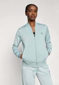 Jasnoniebieska kurtka na zamek wykonana z gładkiego materiału, z kołnierzem typu stójka i bocznymi kieszeniami, w zestawie z dopasowanymi jasnoniebieskimi spodniami.