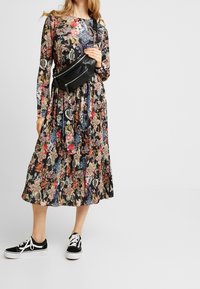 Blumenmuster Kleid mit langen Ärmeln, schwarzem Hintergrund, plissiertem Design und kräftigen Farben. Kombiniert mit einer schwarzen Bauchtasche und Sneakers.