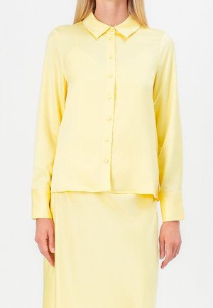 Femme portant une chemise boutonnée à manches longues jaune pâle avec col et une jupe assortie sur fond blanc.