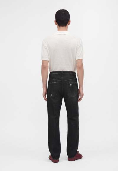 Homme debout de dos, portant un polo blanc à manches courtes, un jean noir avec une légère usure sur les poches arrière et des baskets rouges.