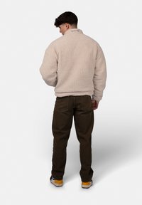 Beige Fleece-Pullover mit hohem Kragen, strukturiertem Oberflächen und gesticktem Logo auf dem Rücken; kombiniert mit dunkelgrünen Hosen und Sneakers.
