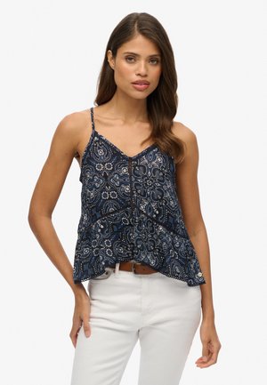 Mujer que lleva una blusa sin mangas de patrón cachemir azul marino con tirantes finos y pantalones blancos, de pie contra un fondo liso.