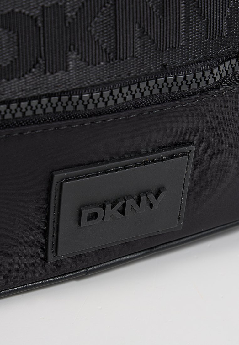 Bolso negro DKNY con tejido de nailon texturizado, que cuenta con una placa de logo negra prominente y un zipper negro. Diseño liso y elegante con bordes redondeados.