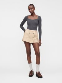 Femme portant un haut gris ajusté à manches longues, une jupe portefeuille beige avec des boutons, des chaussettes blanches et des chaussures compensées marron, debout sur un fond blanc.