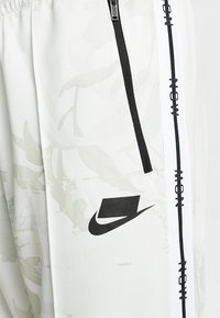 Ljust grå joggers med ett subtilt blommönster, svarta sidostråkar med text och en svart Nike swoosh-logotyp. Fickan är zip-försedd.