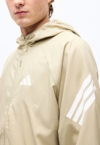 Νεαρός άνδρας που φοράει μπεζ μπουφάν με κουκούλα με λευκές διαγώνιες ρίγες στο στήθος και στο μανίκι, απεικονίζεται από τους ώμους μέχρι το μέσο του προσώπου.