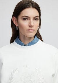 Sudadera blanca con un diseño bordado que presenta una corona y hojas, superpuesta sobre una camisa de denim con un cuello clásico.