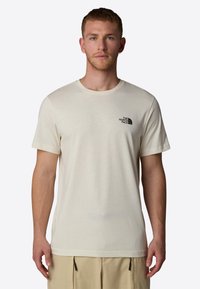 Krémové tričko s krátkým rukávem vyrobené z měkké tkaniny, s malým černým logem na levé části hrudníku a klasickým kulatým výstřihem.