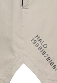 Beżowa tkanina z czarnym nadrukiem tekstu "HALO 1968|972|981" i widocznymi detalami przeszyć w pobliżu krawędzi kieszeni.