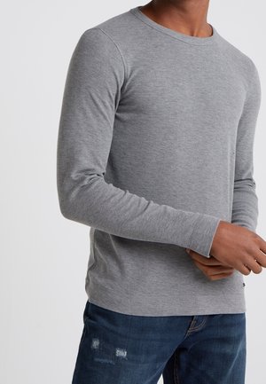 Mann trägt einen eng anliegenden grauen Langarmpullover mit Rundhalsausschnitt und dunkelblaue, abgenutzte Jeans vor einem schlichten Hintergrund.